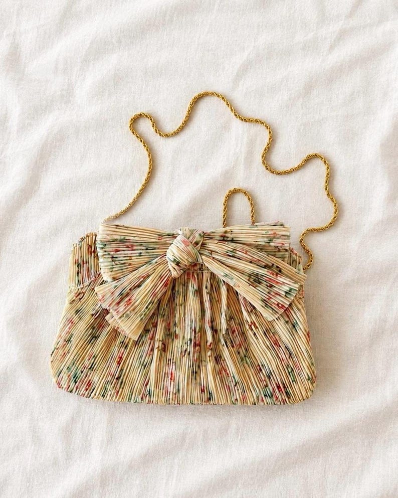Loeffler Randall Rayne Tan Vine Floral pleated bow clutch with gold chain strap on white linen background.