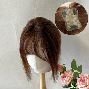 May include: A brown hair topper with a fringe, displayed on a white mannequin head. The topper has a natural look, with varying shades of brown. A close-up shows the base with clips for attachment. The background is a soft, neutral color.