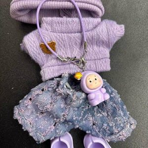 Puede incluir: Un conjunto en miniatura de color lavanda para mu&ntilde;eca, que incluye gorro, su&eacute;ter, pantalones cortos de mezclilla desgastados y zapatos. El su&eacute;ter tiene un peque&ntilde;o parche de oso. Un dije con una figura sonriente cuelga de un collar. El gorro tiene una etiqueta "Hand made".
