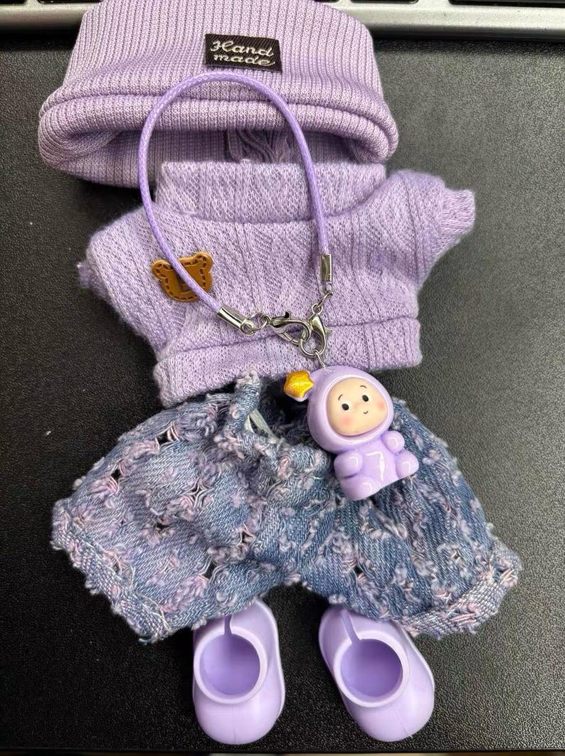Puede incluir: Un conjunto en miniatura de color lavanda para mu&ntilde;eca, que incluye gorro, su&eacute;ter, pantalones cortos de mezclilla desgastados y zapatos. El su&eacute;ter tiene un peque&ntilde;o parche de oso. Un dije con una figura sonriente cuelga de un collar. El gorro tiene una etiqueta "Hand made".