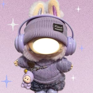 Puede incluir: Un peluche vestido con un su&eacute;ter morado, pantalones cortos de mezclilla y botas, con un gorro de punto y auriculares. El juguete tiene orejas de conejo y un peque&ntilde;o amuleto. El gorro tiene una etiqueta "Hand made".