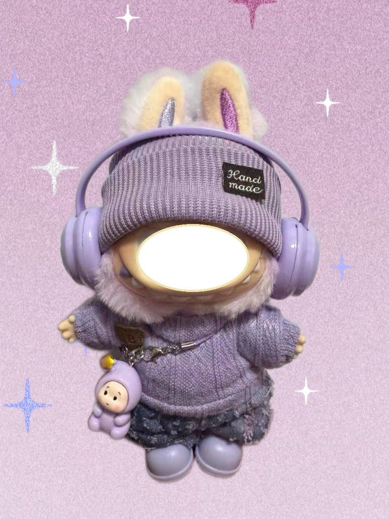 Puede incluir: Un peluche vestido con un su&eacute;ter morado, pantalones cortos de mezclilla y botas, con un gorro de punto y auriculares. El juguete tiene orejas de conejo y un peque&ntilde;o amuleto. El gorro tiene una etiqueta "Hand made".