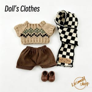 May include: Doll's clothes set featuring a beige sweater with a green and black pattern, brown corduroy shorts, a black and white checkered scarf, and brown doll shoes. The set includes a small tag with a bear illustration.