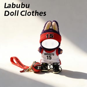 Puede incluir: Un muñeco de peluche con forma de conejo que lleva una diadema roja con el número 15, una camiseta de baloncesto blanca y roja con "Nuggets 15" y zapatillas negras. El muñeco tiene un llavero de baloncesto y el texto "Labubu Doll Clothes".