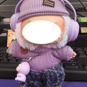 Puede incluir: Mu&ntilde;eca de peluche con gorro de punto morado con orejas de conejo, auriculares a juego y un su&eacute;ter. La mu&ntilde;eca tiene pantalones vaqueros y una peque&ntilde;a figura morada adjunta. El gorro tiene una etiqueta "Hand made".