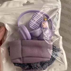 Puede incluir: Un conjunto de accesorios de mu&ntilde;eca en miniatura, que incluye unos auriculares morados, un gorro morado y unos pantalones cortos vaqueros. Los auriculares tienen un dise&ntilde;o a rayas en la parte superior. El conjunto est&aacute; empaquetado en una bolsa de pl&aacute;stico transparente.
