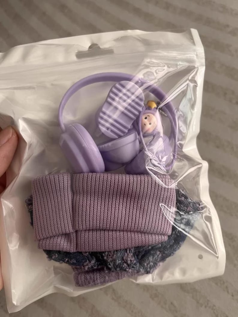 Puede incluir: Un conjunto de accesorios de mu&ntilde;eca en miniatura, que incluye unos auriculares morados, un gorro morado y unos pantalones cortos vaqueros. Los auriculares tienen un dise&ntilde;o a rayas en la parte superior. El conjunto est&aacute; empaquetado en una bolsa de pl&aacute;stico transparente.