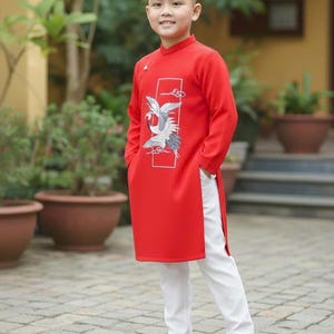 Pu&ograve; includere: Un giovane indossa una tunica rossa con un disegno di uccello bianco e grigio, abbinata a pantaloni bianchi e sandali neri. L'outfit &egrave; in stile tradizionale, adatto per eventi culturali. Lo sfondo comprende vegetazione e piante in vaso.