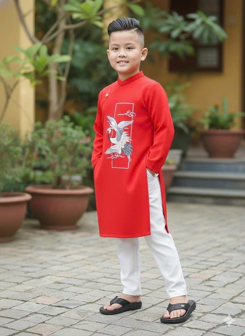 Pu&ograve; includere: Un giovane indossa una tunica rossa con un disegno di uccello bianco e grigio, abbinata a pantaloni bianchi e sandali neri. L'outfit &egrave; in stile tradizionale, adatto per eventi culturali. Lo sfondo comprende vegetazione e piante in vaso.