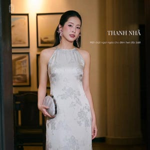 May include: A sleeveless, floor-length, light gray dress featuring a subtle tonal floral pattern, a high halter neckline, and a slim silhouette. The garment is paired with a small, rounded, pleated clutch bag with a metallic frame and clasp.