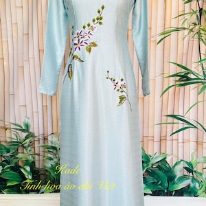May include: A light blue, long-sleeved dress with floral embroidery. The dress features a mandarin collar and a flowing silhouette. The text "Kade Tinh hoa ao dai Viet" is visible on the dress. The dress is displayed on a mannequin.