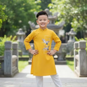 Pu&ograve; includere: Un bambino indossa una tunica giallo senape con un disegno di uccelli e pantaloni bianchi, abbinati a sandali marroni. Lo sfondo presenta un sentiero in pietra e vegetazione verde. L'outfit &egrave; tradizionale.