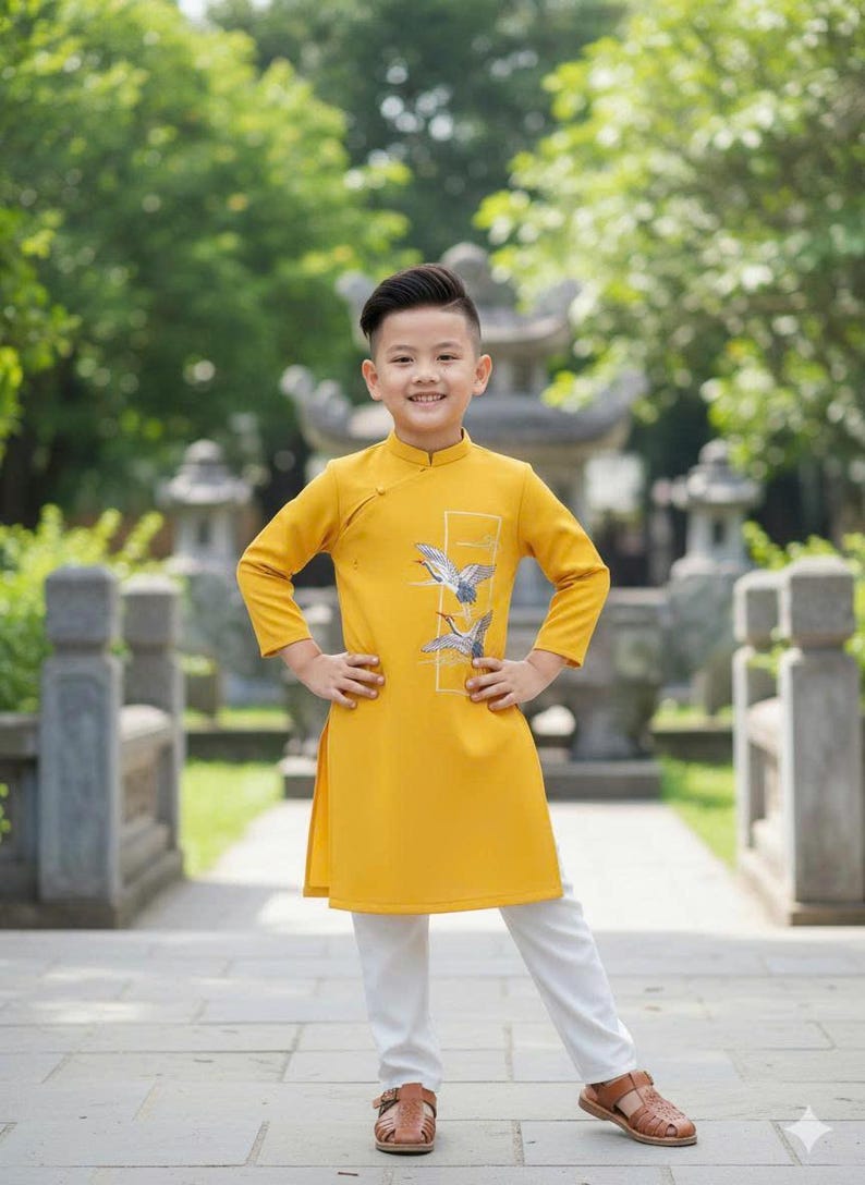 Pu&ograve; includere: Un bambino indossa una tunica giallo senape con un disegno di uccelli e pantaloni bianchi, abbinati a sandali marroni. Lo sfondo presenta un sentiero in pietra e vegetazione verde. L'outfit &egrave; tradizionale.