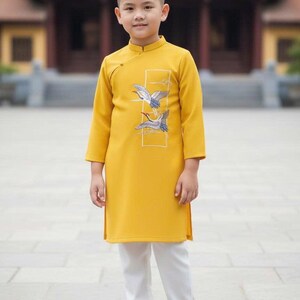 Pu&ograve; includere: Una persona giovane indossa una tunica giallo senape con colletto alla coreana e maniche lunghe, abbinata a pantaloni bianchi e sandali marroni. La tunica presenta un disegno ricamato di due uccelli. Lo sfondo mostra un edificio tradizionale.