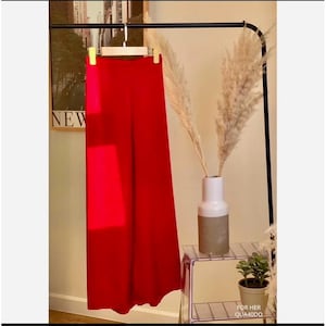 May include: A vibrant red skirt with a smooth, solid-colored fabric hanging from a white hanger on a black metal rack. The skirt features a straight, floor-length cut with a clean hemline, showcasing a bright, saturated hue and a simple design.