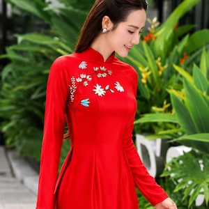 May include: A vibrant red dress with long sleeves and a mandarin collar. The dress features delicate floral embroidery in white and blue near the neckline. The woman is wearing pearl earrings. The background is a lush green garden.