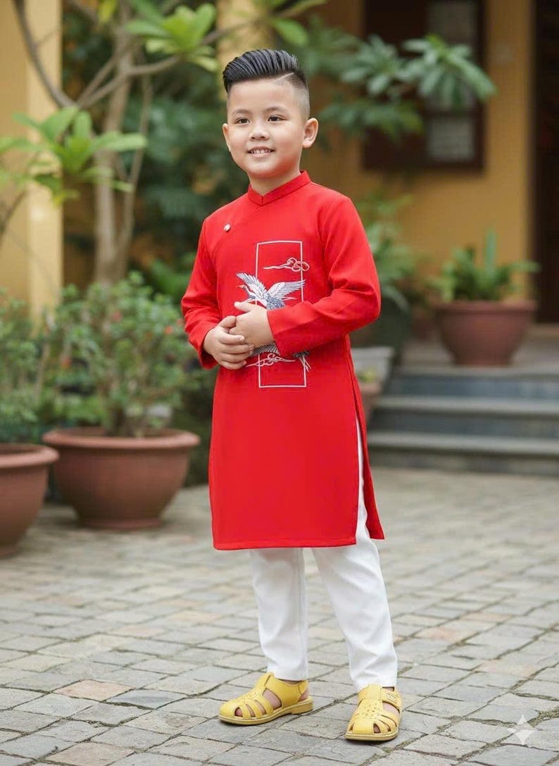 Pu&ograve; includere: Una persona giovane indossa una tunica rossa con un disegno di gru bianca e pantaloni bianchi. L'outfit &egrave; completato da sandali gialli. La tunica ha un colletto alla coreana e maniche lunghe. La persona &egrave; in piedi su un patio di mattoni.