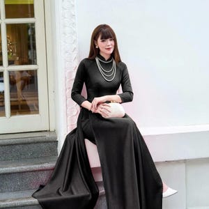 May include: A black long-sleeved Ao dai dress with a high collar and side slits, paired with matching black trousers. The garment features a smooth, solid fabric finish. A multi-strand pearl necklace and a small white clutch are worn with the outfit.