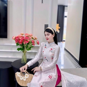 May include: A woman wearing a white, long-sleeved dress with red floral embroidery. The dress has a high collar and a long train. She is holding a small, woven handbag decorated with white flowers. A vase of pink roses sits on a black table.