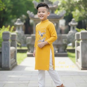 Pu&ograve; includere: Un giovane indossa una tunica giallo senape con un disegno di gru, abbinata a pantaloni bianchi e sandali marroni. L'outfit &egrave; in stile tradizionale vietnamita, con il testo "Kade Tinh hoa ao dai Viet".