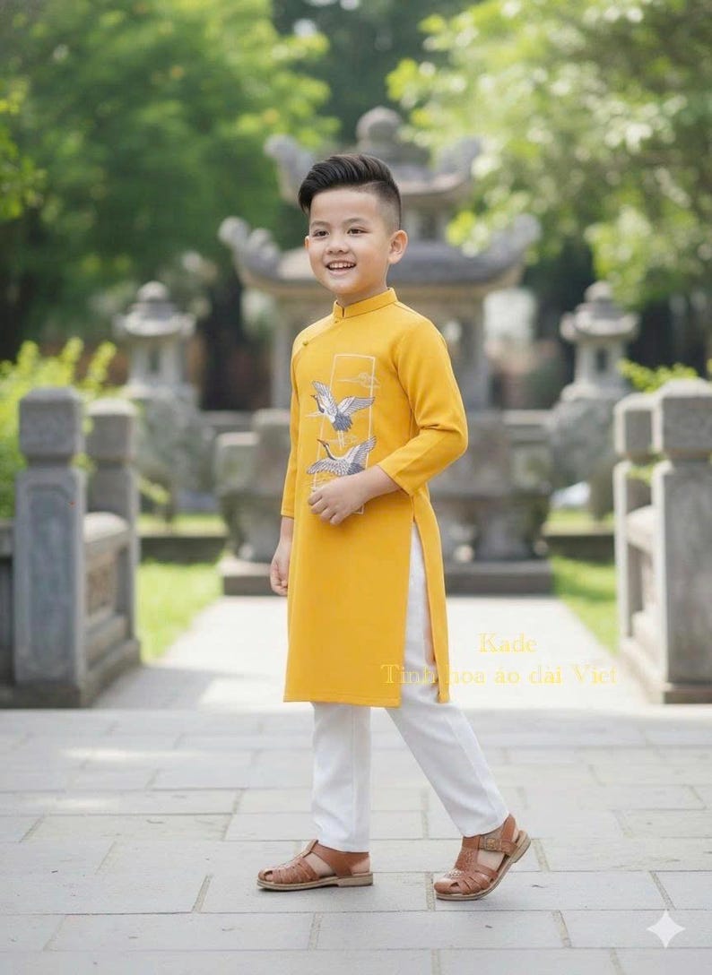 Pu&ograve; includere: Un giovane indossa una tunica giallo senape con un disegno di gru, abbinata a pantaloni bianchi e sandali marroni. L'outfit &egrave; in stile tradizionale vietnamita, con il testo "Kade Tinh hoa ao dai Viet".