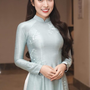 May include: A light blue, long-sleeved dress with floral embroidery. The dress has a mandarin collar and a fitted silhouette. The woman is smiling, with long dark hair. The dress appears to be made of a silky material.