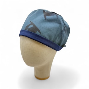 May include: A reversible cap in blue and navy, patterned with black orca whales. The cap has a navy blue brim and is displayed on a mannequin head. The cap is made of a soft fabric.