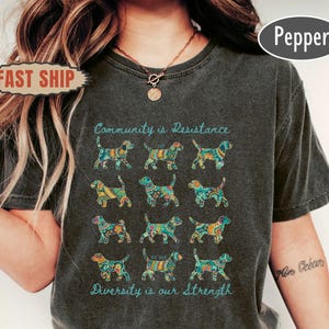 May include: Dark grey t-shirt with a repeating pattern of colourful dog illustrations. The text "Community is Resistance" and "Diversity is our Strength" are printed above and below the dog pattern.
