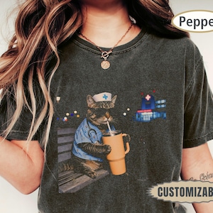 May include: A charcoal gray t-shirt featuring a graphic of a tabby cat dressed as a nurse, wearing a blue scrub top, a stethoscope, and a nurse cap, while holding a large orange tumbler with a straw. A hospital building icon is visible nearby.