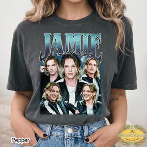 Op de afbeelding: Een antracietgrijs T-shirt met korte mouwen en een grafische print van Jamie Campbell Bower. Het ontwerp bevat de naam JAMIE in grote groenblauwe en witte letters, de tekst Campbell Bower en vier verschillende portretfoto's van de acteur.