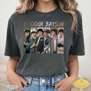 May include: A charcoal gray short-sleeve t-shirt featuring the text MADDOX BATSON in a multi-colored font above six vertical panels. Each panel displays a different photograph of the same person wearing various outfits, including jackets and a cap.
