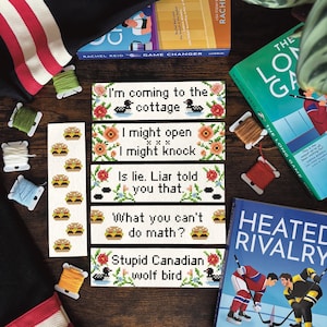 May include: Handmade cross-stitch bookmarks with funny quotes and burger patterns. The image includes spools of thread and books, such as "The Long Game" and "Heated Rivalry", on a wooden surface.