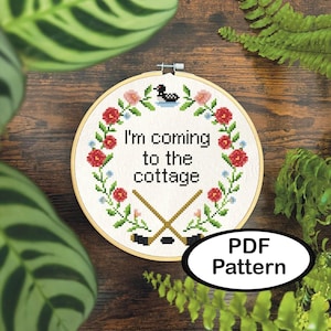 May include: Cross-stitch embroidery hoop with the text "I'm coming to the cottage" surrounded by a floral wreath. The design includes crossed hockey sticks and a small duck. A white oval with "PDF Pattern" is in the lower right.