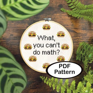 Puede incluir: Un bastidor de bordado de punto de cruz con un diseño de iconos de hamburguesas pixeladas y el texto "What, you can't do math?" También se incluyen las palabras "PDF Pattern". El bastidor está sobre un fondo de madera con follaje verde.