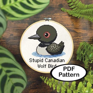 May include: A cross-stitch pattern of a cartoon loon bird in a wooden embroidery hoop. The bird is black, white, and gray with a red eye, floating on blue water. Text reads "Stupid Canadian Wolf Bird" and "PDF Pattern".