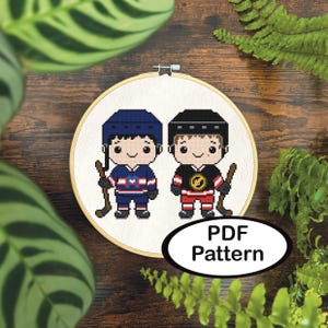 Op de afbeelding: Kruissteekpatroon met twee cartoon hockey spelers in blauwe en zwarte uniformen, elk met een hockeystick. Het ontwerp staat in een houten borduurring, met de tekst "PDF Pattern" in een ovaal. De achtergrond bevat groen gebladerte.