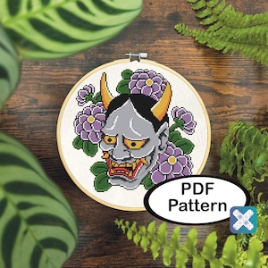 May include: Cross-stitch pattern of a gray demon face with yellow horns and eyes, surrounded by purple flowers and green leaves, set in a wooden embroidery hoop. The text "PDF Pattern" is visible.