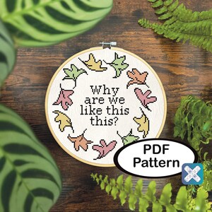 May include: A cross-stitch hoop with the text "Why are we like this?" surrounded by colorful leaf designs. The PDF pattern is displayed on a wooden surface, framed by green foliage.