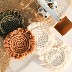 May include: A collection of handmade macrame coasters in various colors, including olive green, tan, and cream. The coasters have a spiral design with fringed edges, perfect for protecting surfaces from drink condensation.
