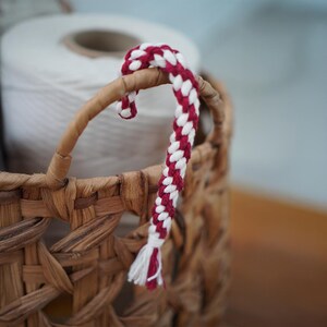 May include: A red and white braided rope handle is attached to a woven basket. The handle is curved and has a tassel at the end. A roll of white thread is in the background.
