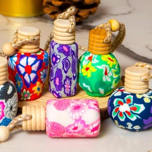 Może przedstawiać: Kolekcja kolorowych, wzorzystych odświeżaczy powietrza do samochodu. Wzory obejmują kwiatowe i abstrakcyjne motywy w różnych kolorach, z drewnianymi nakładkami i sznurkami do zawieszania. Kształty są okrągłe i prostokątne.