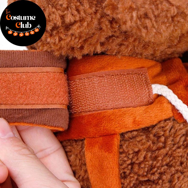 May include: Close-up of a brown and orange costume with a hook and loop fastener. The Costume Club logo is in the upper left corner. The costume appears to be made of a soft, plush material.