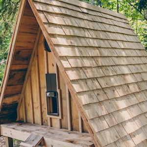 May include: A-frame wooden playhouse with a shingled roof. The structure features a small window and is set in a wooded area. The playhouse is constructed from light-colored wood.