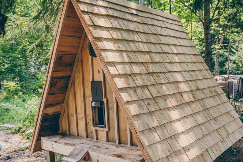 May include: A-frame wooden playhouse with a shingled roof. The structure features a small window and is set in a wooded area. The playhouse is constructed from light-colored wood.
