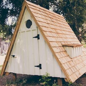 May include: A-frame wooden structure with a white exterior and a cedar shake roof. The structure has a small round window and a small vent. Black hinges and a door latch are visible. The structure is elevated on wooden posts.