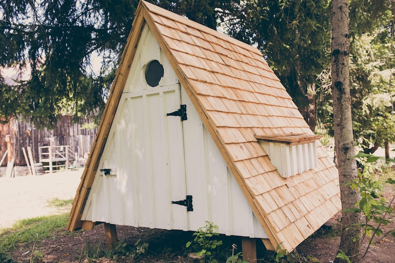 May include: A-frame wooden structure with a white exterior and a cedar shake roof. The structure has a small round window and a small vent. Black hinges and a door latch are visible. The structure is elevated on wooden posts.