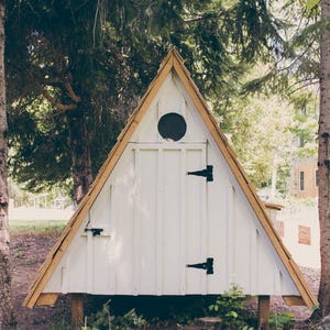May include: A-frame structure, painted white with a natural wood trim and roof. The small building has a circular window and black hinges. It is set in a wooded area with trees and foliage.