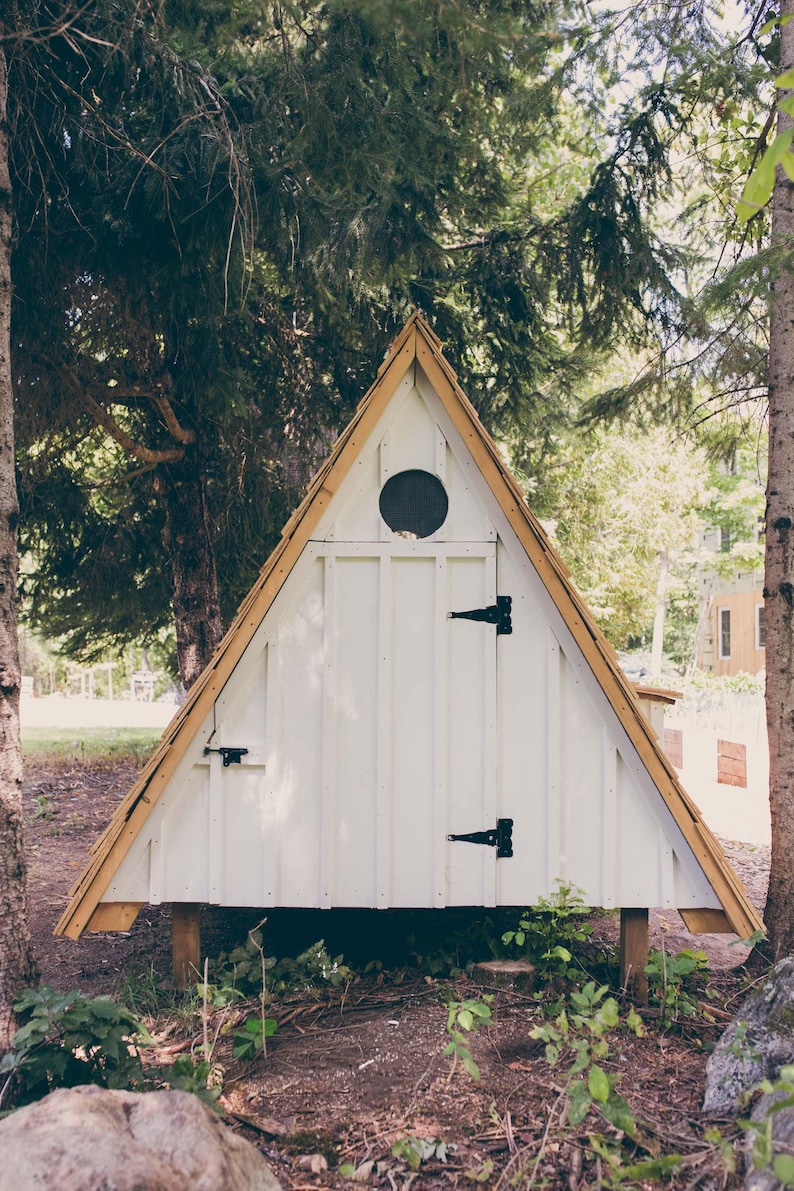 May include: A-frame structure, painted white with a natural wood trim and roof. The small building has a circular window and black hinges. It is set in a wooded area with trees and foliage.