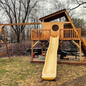 Op de afbeelding: Een houten speelset met een schommel en een speelhuisje met een glijbaan. Het speelhuisje heeft een rond raam en een ladder. De glijbaan is geel. De speelset staat op een grasveld.