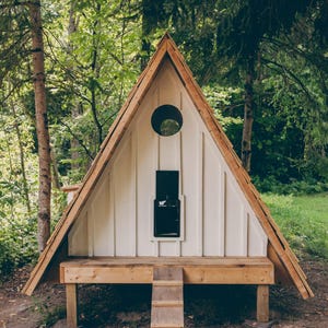 May include: A-frame chicken coop with a white exterior and a wooden roof and trim. The coop has a round window, a black door, and a small wooden ramp leading to a platform. The coop is set in a wooded area.